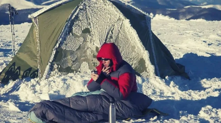 man sitting outside tent in the snow in sleeping bag intext