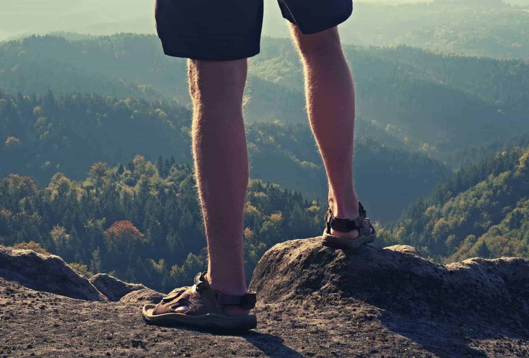 Close up of a mans legs standing on top of a hill wearing hiking sandals