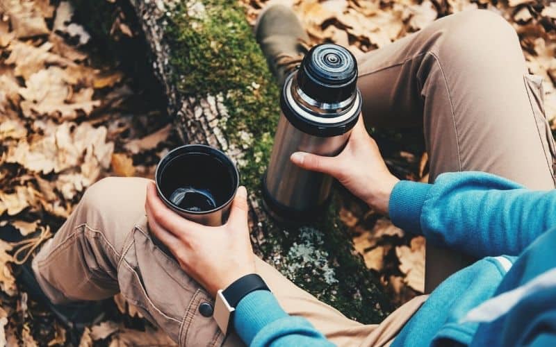 camper drinking from thermos flask sitting on log