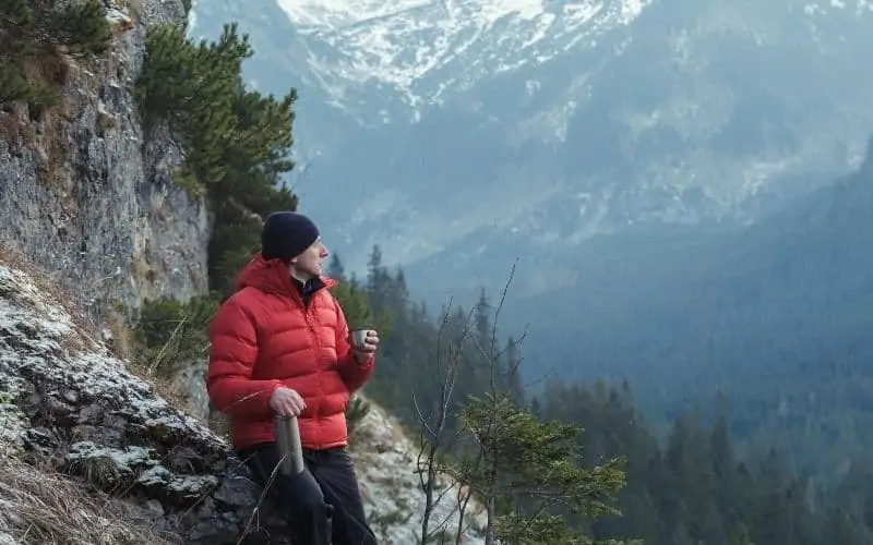 hiker in down jacket drinking from thermos overlooking mountain valley