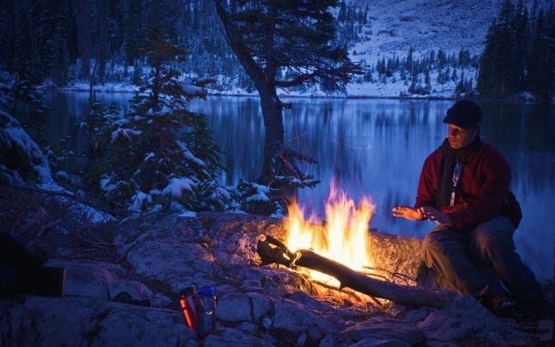 Man in snowy lakeside scene warming hands by campfire