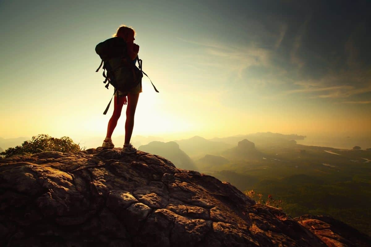 Woman with bacpack on stands on mountaintop looking out at the view