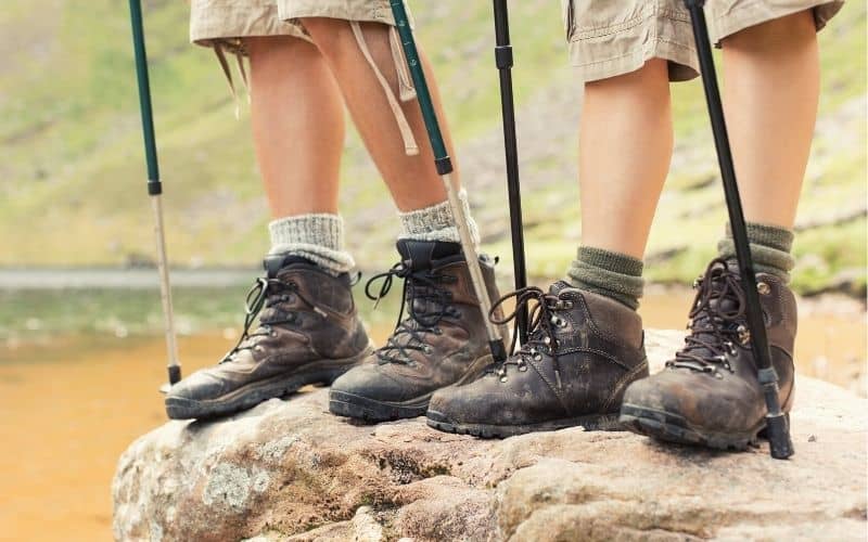 Close up of two peoples legs wearing hiking boots and standing on a rock holding trekking poles