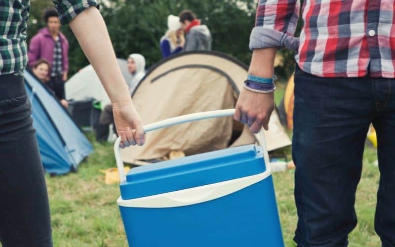 Two people carry a hard walled cooler toward camp