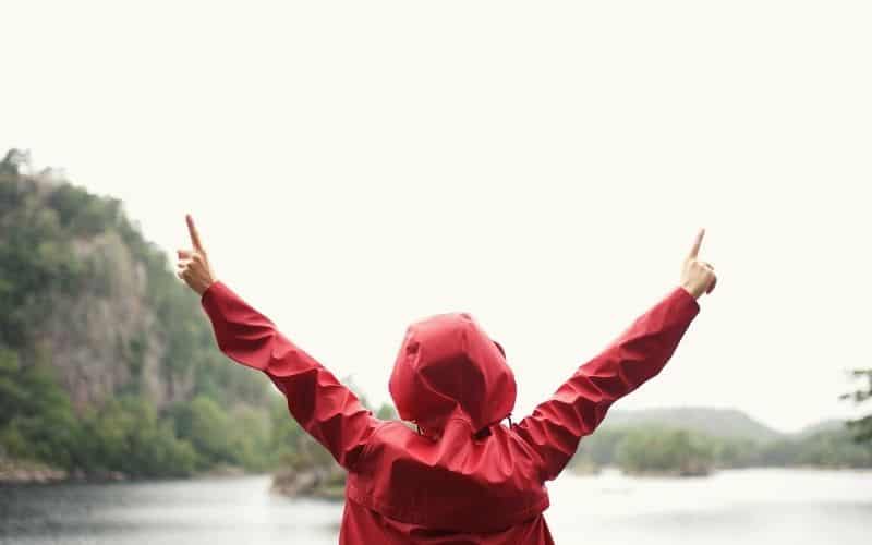 Woman wearing red rain jacket with hands in air looking over a lake