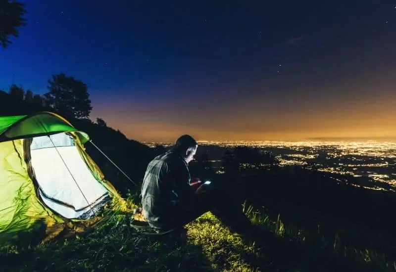 Man sitting on a mountaintop on his phone and beside his well lit tent
