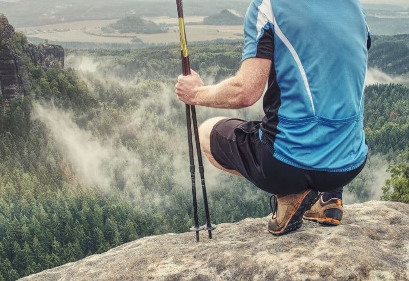 Man crouching on mountaintop wearing t-shirt and holding trekking poles