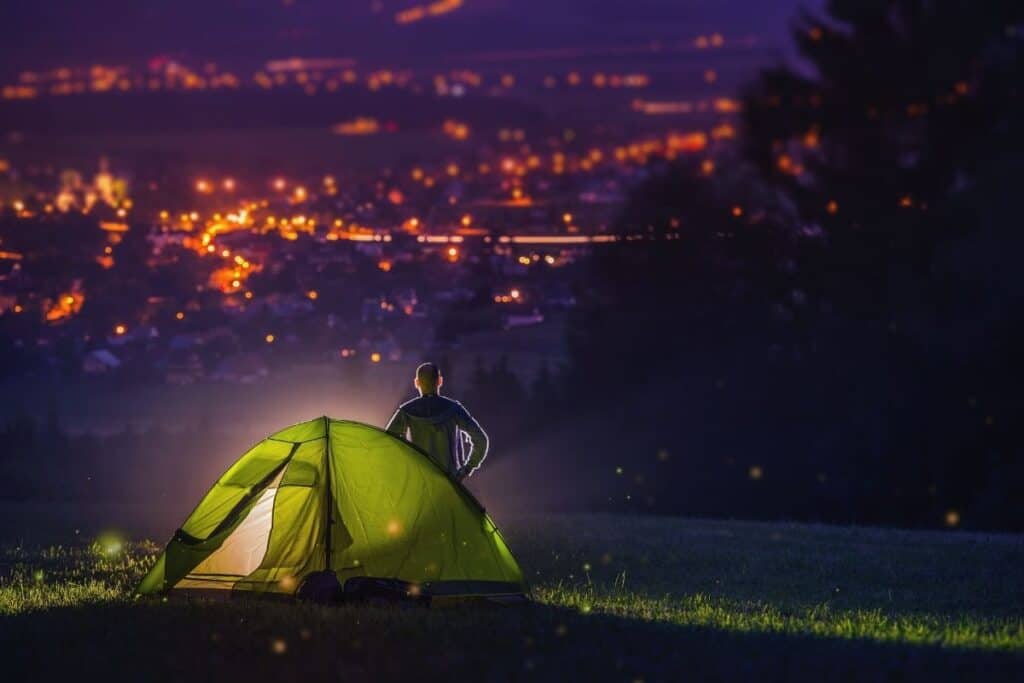 Light shining out of tent pitched on a hilltop overlooking a well lit city