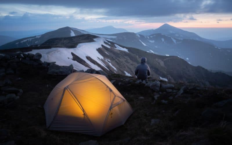 Man sitting outside tent at dusk looking at snowy mountains