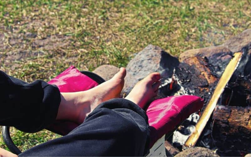 Feet resting on a camping chair footrest in front of a campfire