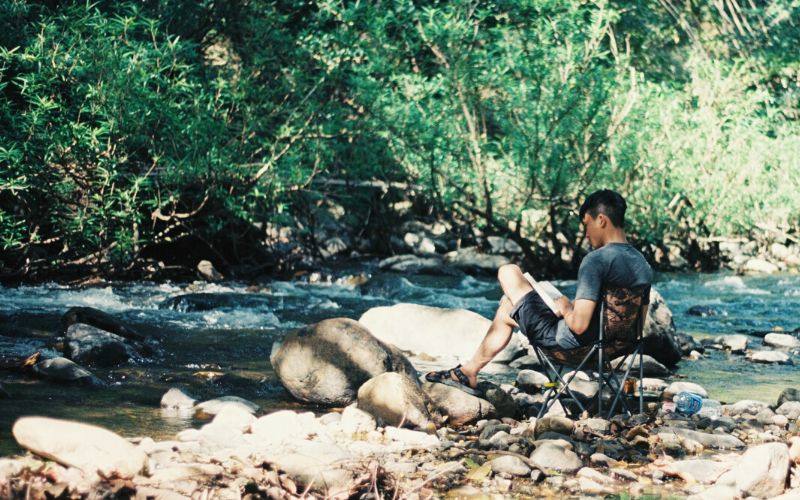 Man sitting in a camp chair reading a book at a riverside