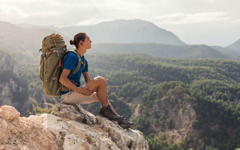 Man wearing backpack sitting on top of rock in front of mountains