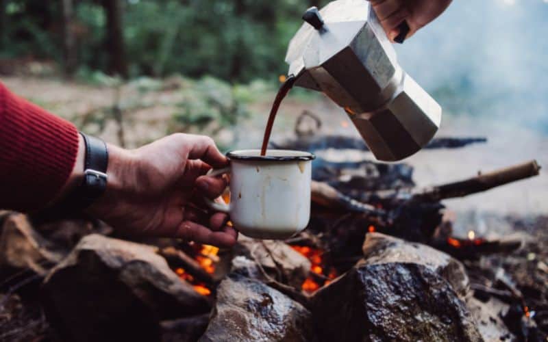 Camper pouring coffee from moka pot into mug over campfire (SS)