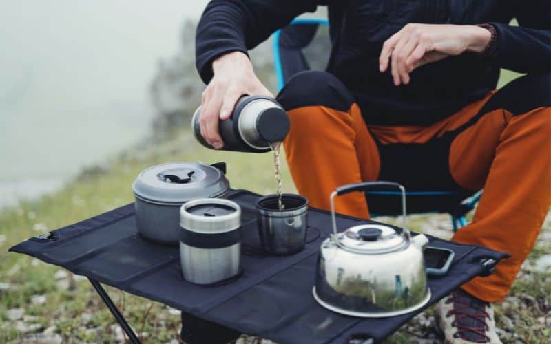 Camper pouring water from thermos into mug on camp table with cooking gear on it