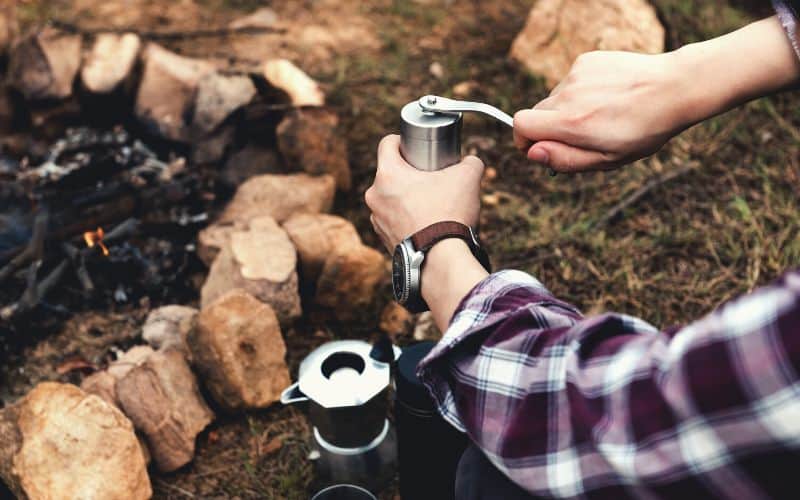 Camper using a coffee grinder to grind coffee beside campfire 