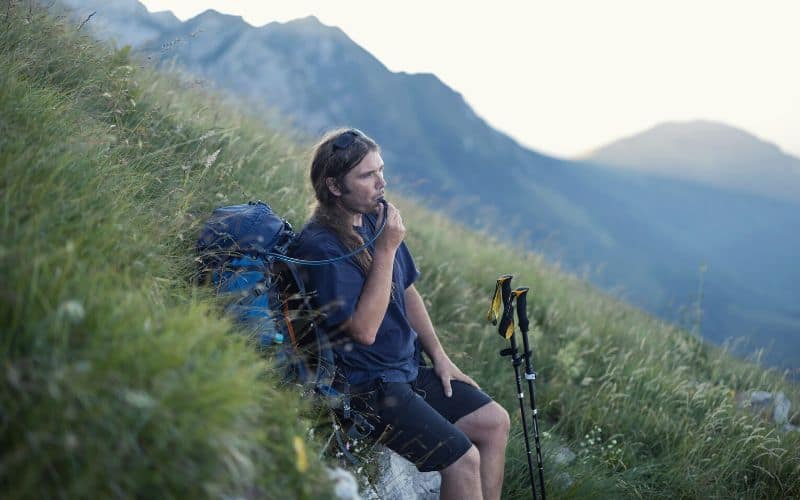 Hiker drinking from camel pack tube with his hiking poles and backpack beside him