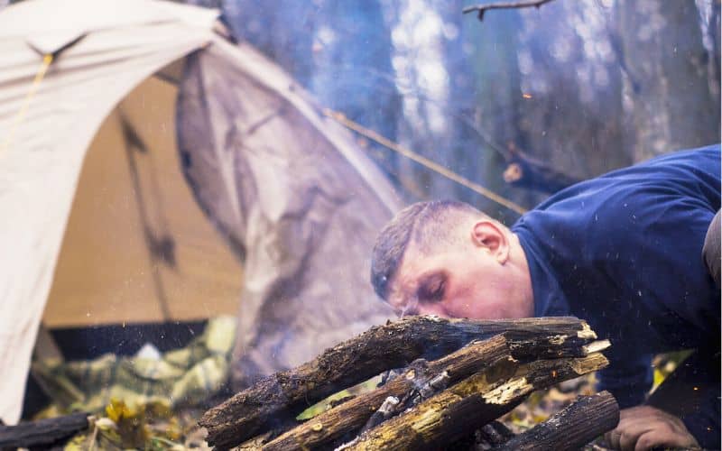 Man blowing on tinder under a pile of logs with smoke coming out
