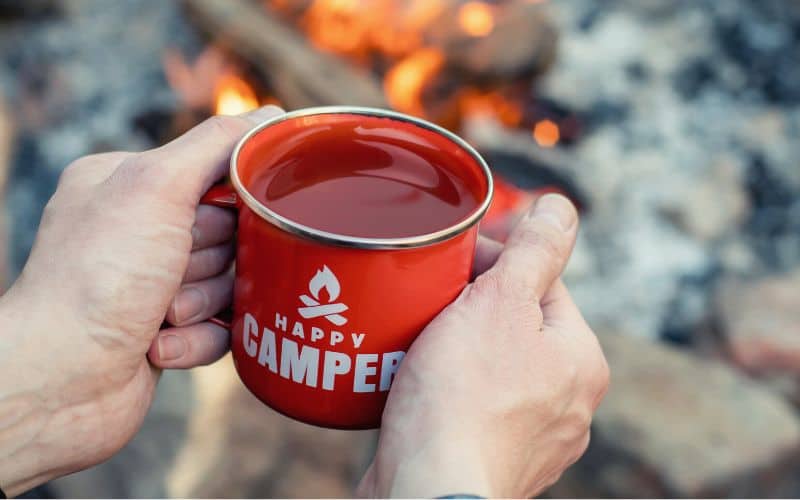 Person holding a mug that says happy camper on it in front of a campfire 