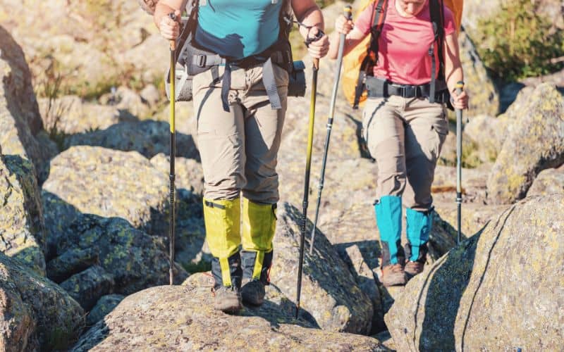 Two hikers hiking over boulders with trekking poles and wearing gaitors 