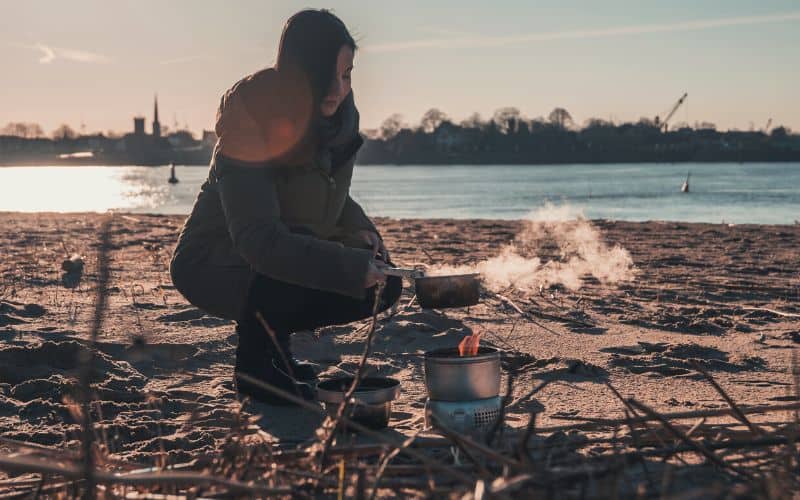Woman boiling water over camping stove in front of a river