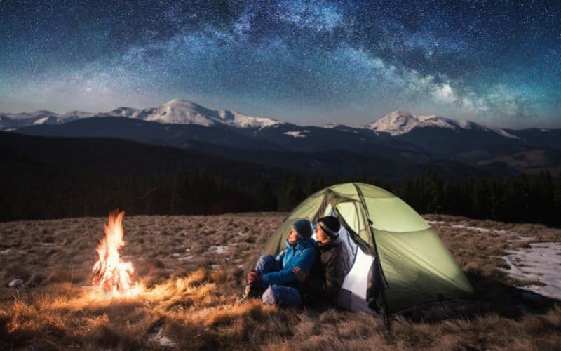 Campers sitting at tent watching campfire and starry night sky 