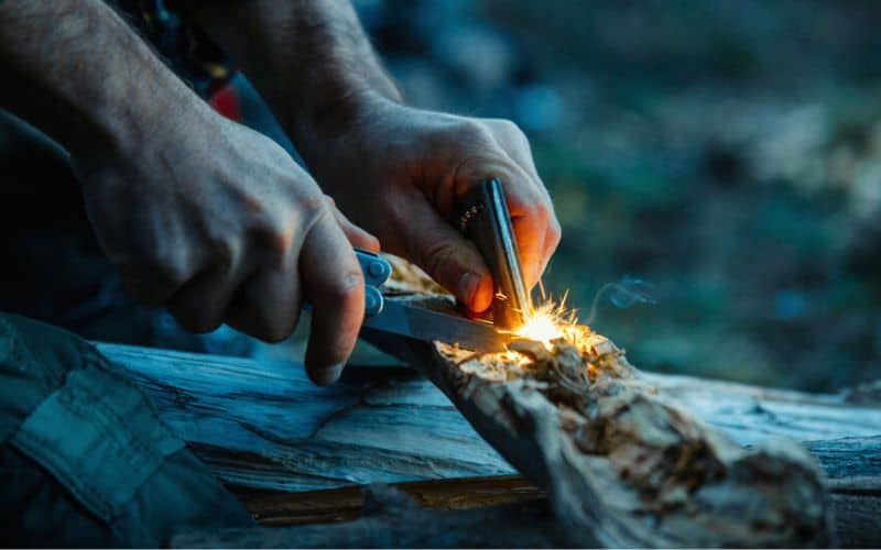 Man at dusk striking a magnesium bar top create sparks over kindling