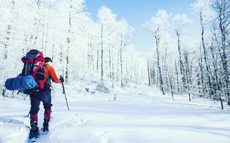 Man hiking through snow wearing gaitors, a backpack with sleeping pad attached and holding trekking poles 