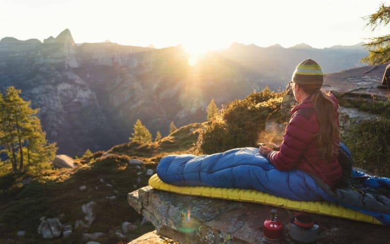 Camper sitting on sleeping pad in front of the sun rising over mountains (SS)