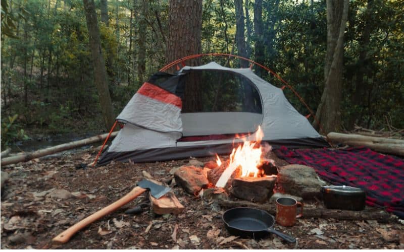 Campfire with frying pan axe and other cooking utensils beside it in front of a tent pitched in the forest