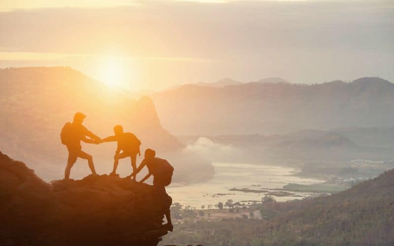 Silhouette of three hikers helping each other to the top of a cliff edge