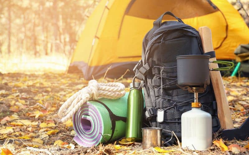 Camping gear including backpack, sleeping mat, and camping stove packed up in front of a tent in the forest