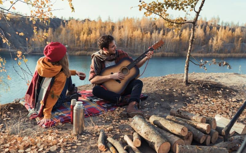 Couple  playing guitar sitting on river bank in front of pile of logs