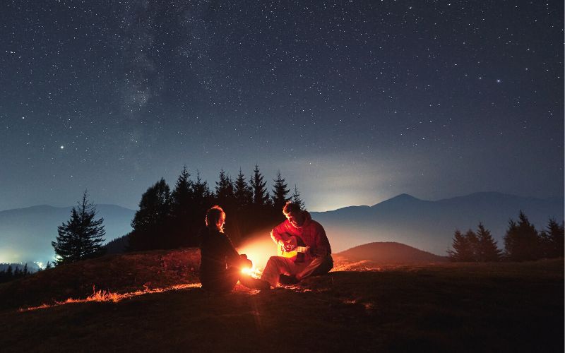 Man playing guitar to woman in front of campfire and under a starry night sky