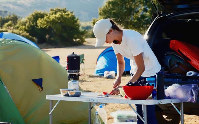 Woman chopping tomatoes on camp table set up beside car trunk 